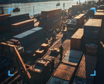 Veduta dall'alto di un porto commerciale con container colorati, operatori con caschi protettivi e un camion, illuminati dalla luce calda del sole.