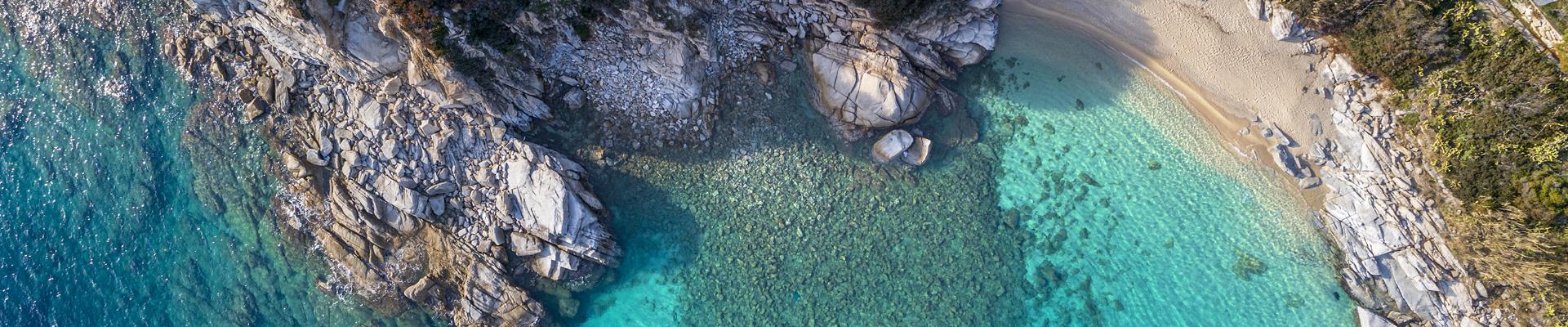 Vista aerea dall'alto di una scogliera rocciosa e una spiaggia dell'Isola d'Elba con mare cristallino dalle sfumature turchesi.